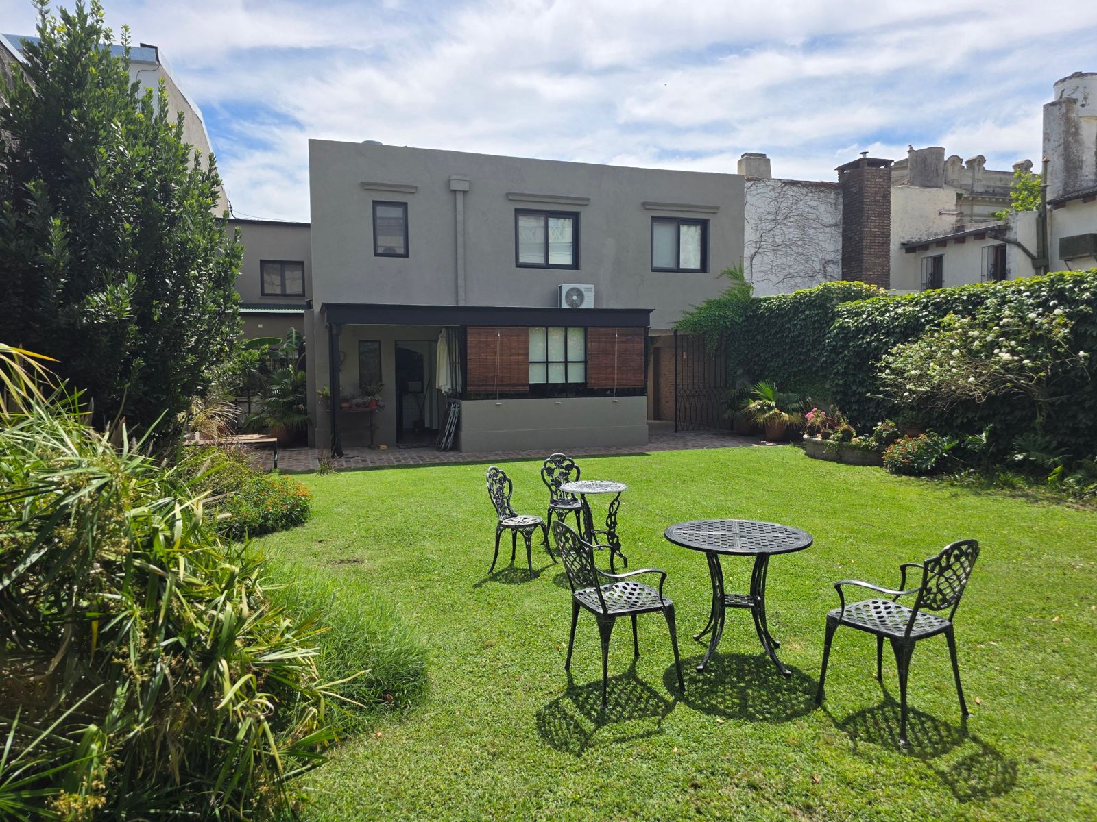 La casa vista desde el jardín, con la galería, el césped y las mesas de hierro